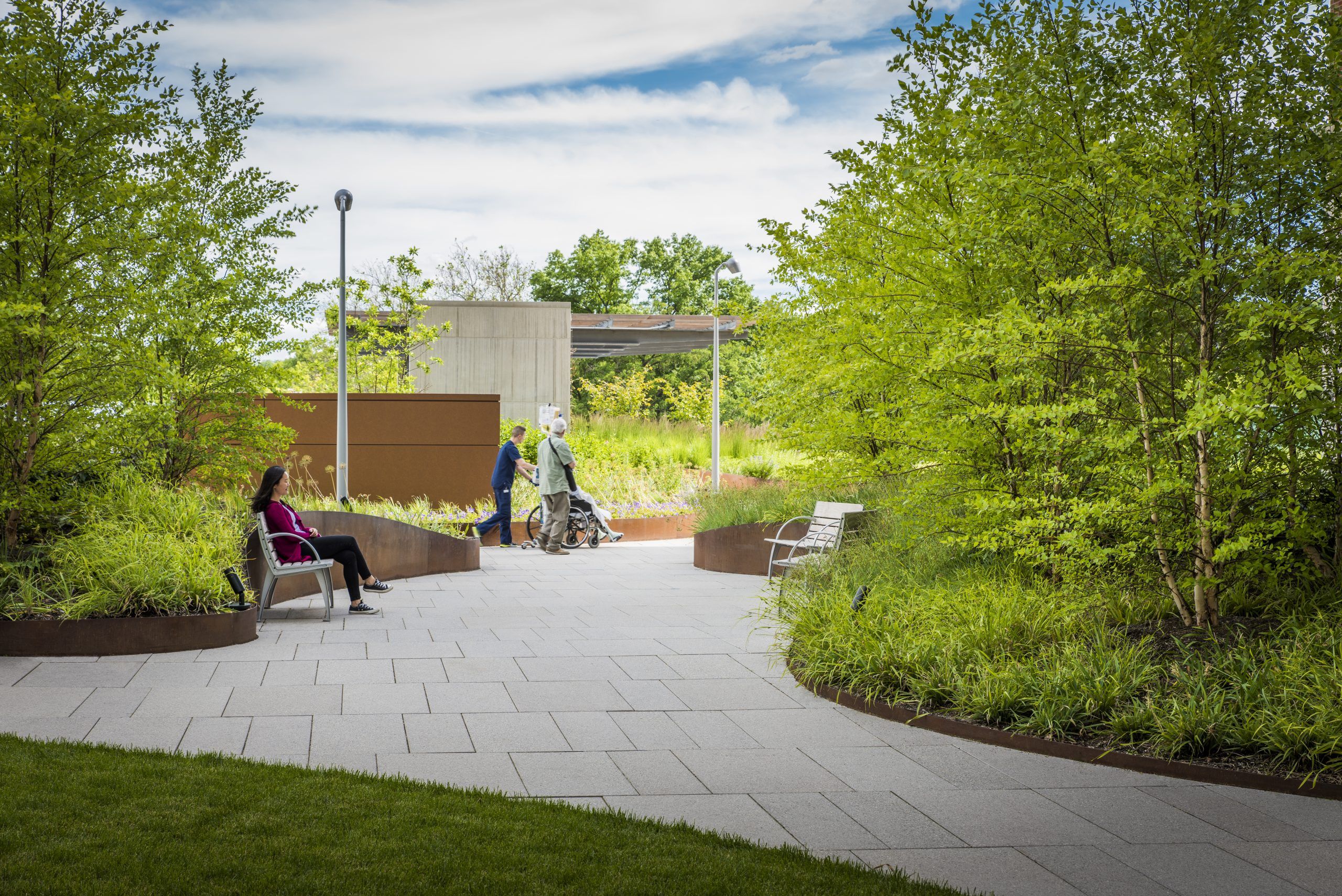 Nurse pushes patient in wheelchair through lit garden courtyard with smooth pavestone path and public seating