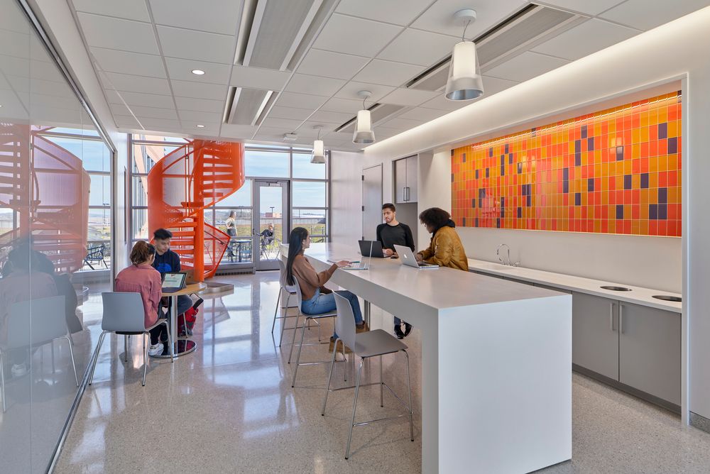 Researchers and students collaborating with laptops in the foreground of a white commons area with a DNA-inspired orange staircase in the background