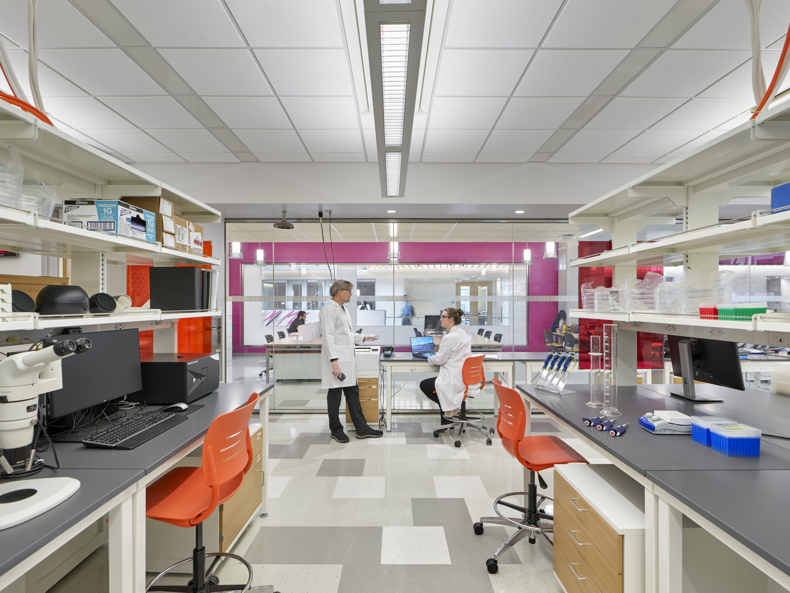 Two researchers talking in a wet lab with patterned, gray-tiled floors, recessed overhead lighting, and workstations with orange chairs