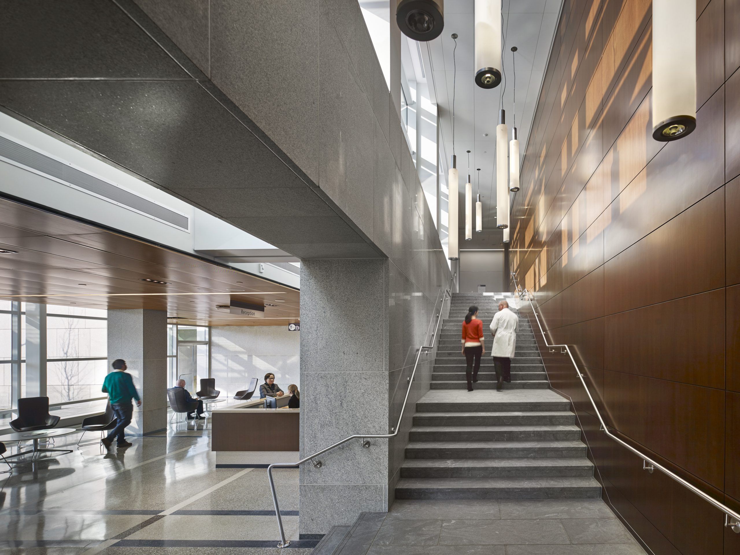 Doctor and patient walking up a gray stone stairway with pendular lighting, which leads from the first-floor reception area to the second floor