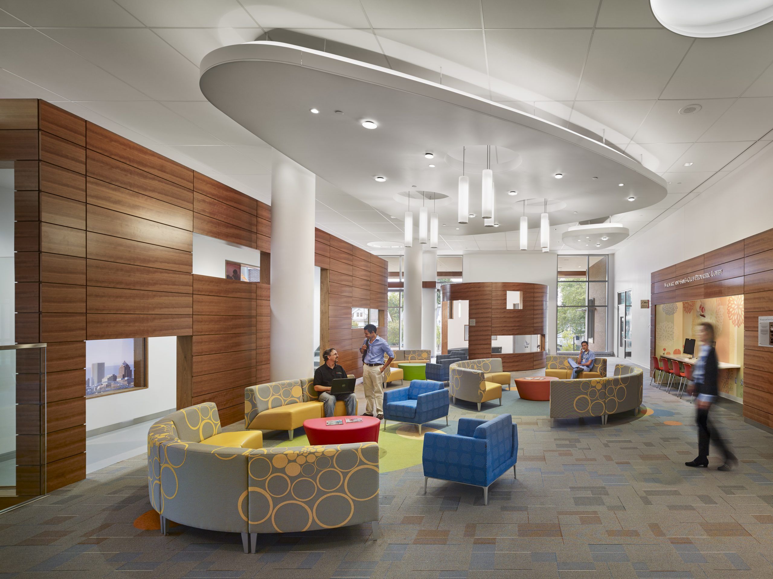 People lounge in a pediatric lobby with wood paneling, colorful furniture, and scaled entrances for children at play