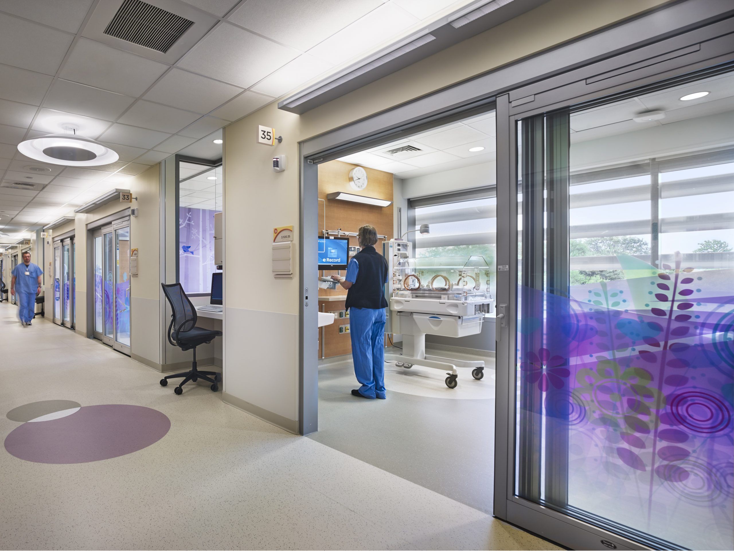 A hospital staff member works in a neonatal room while another nurse walks by a nurses' station outside